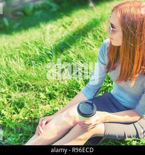 Beautiful red-haired girl sitting on the grass and drinking coffee from a paper Cup. Lifestyle Breakfast in the Park Stock Photo
