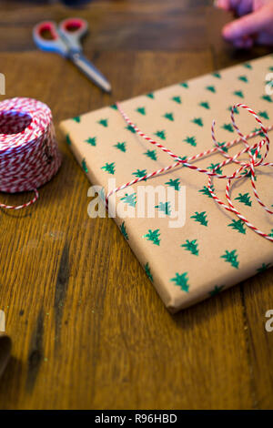 close up of christmas wrapping session on old wooden pine table with parcel in Xmas tree paper, red and white cord. Scissors and a hand in background Stock Photo