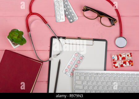 Doctor workplace with blank clipboard, medicine red stethoscope, computer keyboard, eyeglasses, book and pills on pink wooden table. Medication prescr Stock Photo