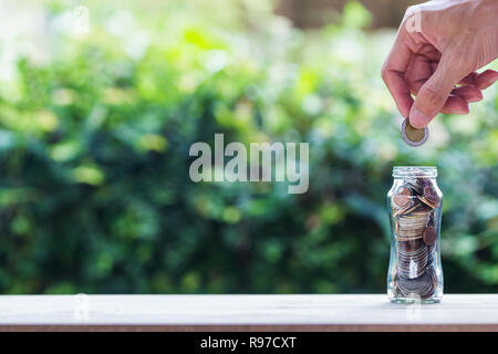 Financial investment / Savings money growing concept : Hand holding coin over coins in glass jar on wood table with nature and space. Conceptual savin Stock Photo