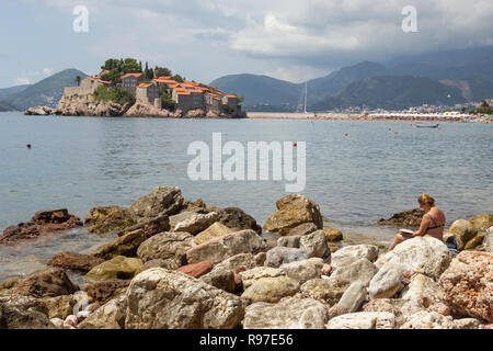 A woman is sitting on the stone and reading a book near the water on the background of the island of St. Stephen on a sunny summer day Stock Photo