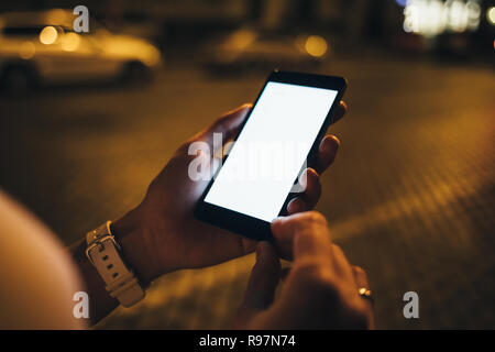 Close-up young woman's hands holding mobile phone with blank white screen outside at street, road and city lights on background. Stock Photo