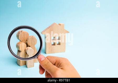 Magnifying glass is looking at the family stands near a wooden house on a blue background. The concept of affordable housing and mortgages for buying  Stock Photo