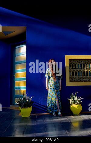 A long haired brunette lady wearing a blue and yellow lemon patterned dress looks upwards, posing on a blue and yellow garden terrace, lit sideways by Stock Photo