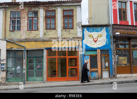 Typical street of Veliko Tarnovo where many business are now closed with cat street art. Old man wearing black and a cane walking up the hill Stock Photo