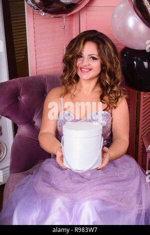 charming smiling curly young woman in a beautiful long dress with a gift on the background of the fireplace, New Year and Christmas decor, balloons Stock Photo