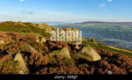 Scenic rural landscape of blue sky & early morning sun on rocky outcrop & mist in valley - Cow & Calf Rocks, Ilkley Moor, West Yorkshire, England, UK. Stock Photo