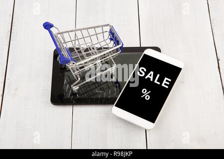 Shopping Cart on the tablet pc near to smartphone with inscription "SALE %" on a white wooden background Stock Photo
