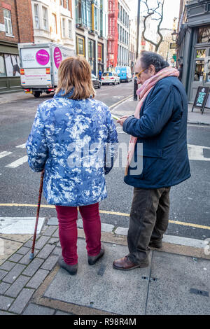 Eccentrically dressed elderly man and woman walking with the aid of a cane discuss crossing a road in London Stock Photo