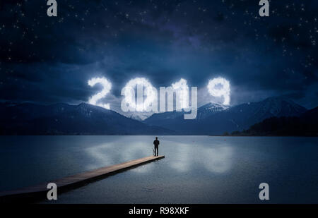 New year background, young man standing on a jetty in a lake and looking to the mountains under the dark sky with cloudy text 2019 Stock Photo