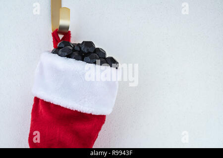 Close up of traditional red and white plush Christmas stocking stuffed with coal shaped candy on a gold hook against a white background Stock Photo
