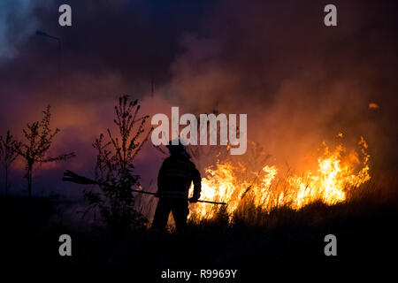 A firefighter fighting fire of bush, using a fire equipement. Some trees and bush burning in the evening. Wide spreaded smoke and flames in the backgr Stock Photo