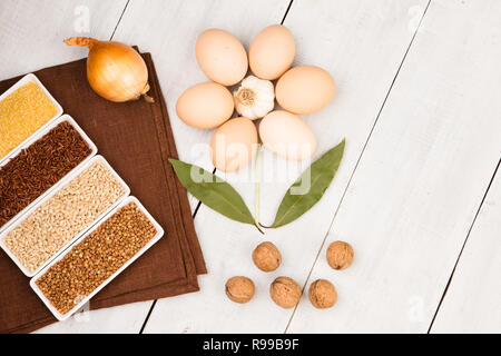 Various groats in bowls, eggs, bay leaf, table-napkin laid out in the shape of a flower on white wooden table Stock Photo