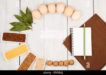 Various groats in bowls, eggs, bay leaf, table-napkin and notepad on white wooden table Stock Photo