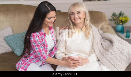 Smiling Daughter Gently Touches Her Mother's Hands. Elegant Senior Woman Sits On The Couch And Holds A Cup Of Tea While Her Young Daughter Hugs Her. Stock Photo