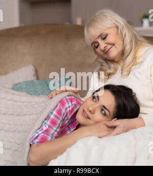 Portrait Of Cute Tired Daughter Lying On Mother's Knees. Senior Mother Is Patting Her Sweet Daughter Who Lays On Her Knees Covered In Warm Blanket Stock Photo