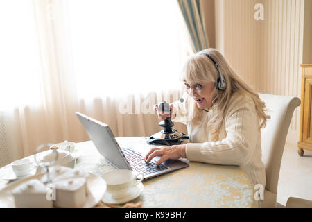 General Shot Of Excited Senior Woman Winning In Video Game Holding A Joystick And Wearing Headphone. Sitting At The Table Playing Games Stock Photo