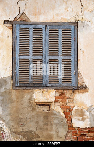 old blue shuttered window in cracked and peeling plaster wall over bricks Stock Photo