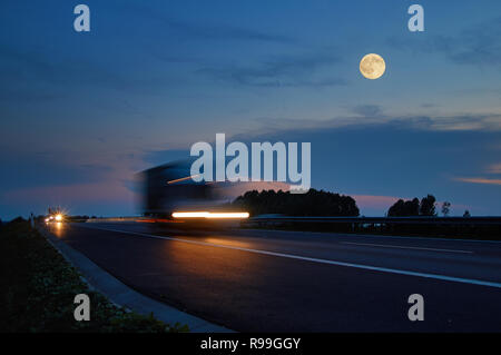 Trucks with glowing lights arriving on the motorway from afar in the landscape with a full moon at dusk. Motion blur speeding trucks. Dark blue summer Stock Photo