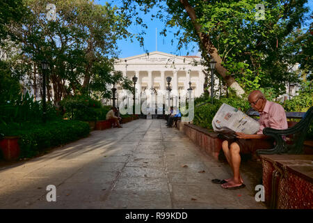 On a late afternoon, a man reads a newspaper in Horniman Circle, Fort area, Mumbai, India; in the backgr. The Library of the Asiatic Society of Mumbai Stock Photo