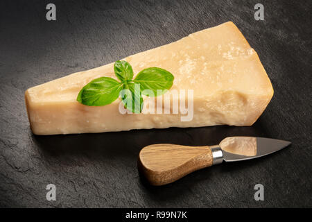 A piece of aged Parmesan cheese on a black background with fresh basil leaves, a cheese knife, and a place for text Stock Photo