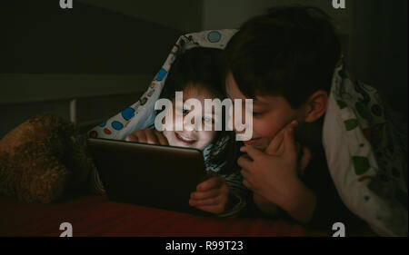 Brother and sister lying on the bed looking at the tablet in the dark Stock Photo