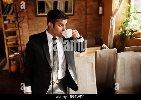 Elegant south asian indian business man in black suit posed indoor cafe and drinking tea. Stock Photo