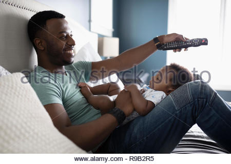 Smiling father with remote control watching TV with baby son in lap on bed