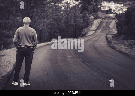 Active senior man standing on lonely road between mountains in black and white. Older man of back standing on lonely highway Stock Photo