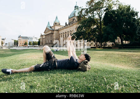 A young guy with a backpack or a student is lying on the grass during a break or is resting and using a tablet. He is watching a video or reading or something else. Stock Photo