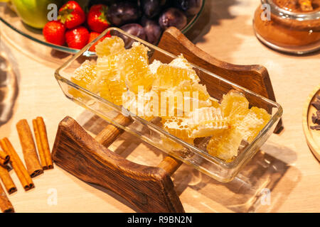 Closeup of fresh honeycombs cut into cubes in transparent square glass bowl at a buffet, cinnamon sticks, fruits, strawberries. Sweets table concept a Stock Photo
