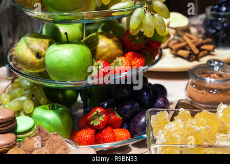 Closeup bright fresh fruits: apples, strawberry, pear, grape, honeycombs cubes in glass bowl, flowers irises, macaroons, truffles on buffet table. Con Stock Photo