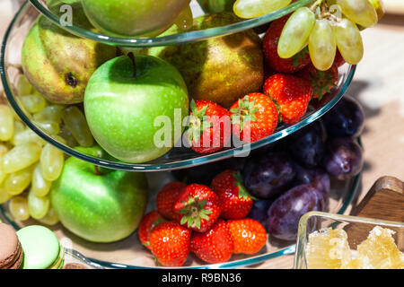 Closeup bright fresh fruits: apples, strawberry, pear, grape, honeycombs cubes in glass bowl, flowers irises, macaroons, truffles on buffet table. Con Stock Photo
