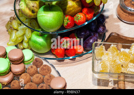 Closeup bright fresh fruits: apples, strawberry, pear, grape, honeycombs cubes in glass bowl, flowers irises, macaroons, truffles on buffet table. Con Stock Photo