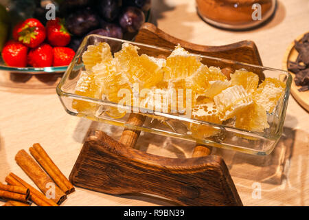Closeup of fresh honeycombs cut into cubes in transparent square glass bowl at a buffet, cinnamon sticks, fruits, strawberries. Sweets table concept a Stock Photo