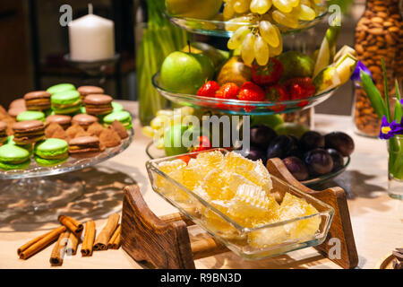 Closeup of fresh honeycombs cut into cubes in transparent square glass bowl at buffet, cinnamon sticks, fruits, macaroons, truffles, nuts, candles. Co Stock Photo