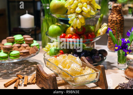 Closeup of fresh honeycombs cut into cubes in transparent square glass bowl at buffet, cinnamon sticks, fruits, macaroons, truffles, nuts, candles. Co Stock Photo