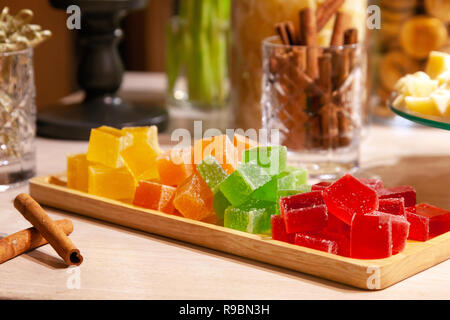 Closeup of stacks of small cubes of green, red, yellow, orange marmalade and cinnamon sticks in crystal glass on rectangular wooden plate on a caterin Stock Photo