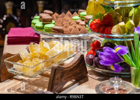 Closeup of fresh honeycombs cut into cubes in transparent square glass bowl at buffet, cinnamon sticks, fruits, macaroons, truffles, nuts, candles. Co Stock Photo