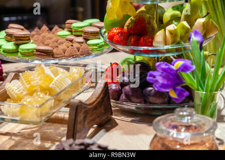 Closeup of fresh honeycombs cut into cubes in transparent square glass bowl at buffet, cinnamon sticks, fruits, macaroons, truffles, nuts, candles. Co Stock Photo