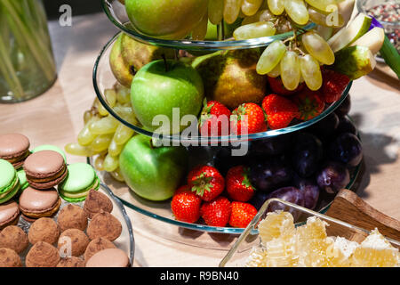 Closeup bright fresh fruits: apples, strawberry, pear, grape, honeycombs cubes in glass bowl, flowers irises, macaroons, truffles on buffet table. Con Stock Photo