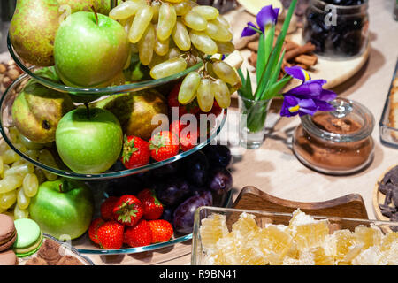 Closeup bright fresh fruits: apples, strawberry, pear, grape, honeycombs cubes in glass bowl, flowers irises, macaroons, truffles on buffet table. Con Stock Photo