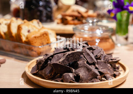 Closeup of pieces of natural dark chocolate on a round wooden plate on the background of sweets, pastries for tasting. Concept diet, healthy food, low Stock Photo