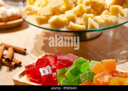 Closeup of stacks of small cubes of green, red, yellow, orange marmalade, glass round plate with hard parmesan cheese and cinnamon sticks on rectangul Stock Photo
