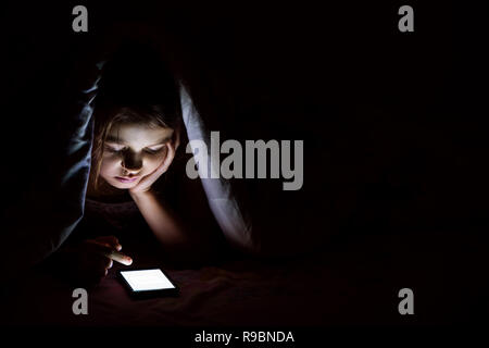 The girl of 9 years old at night is covered with a blanket and is looking into a smartphone. Stock Photo