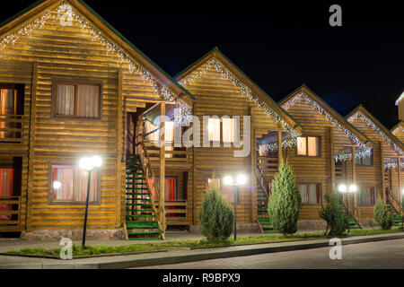 Row of new two-storied ecological wooden comfortable decorated cottages hotels on clean illuminated street under dark night sky. Tourism and recreatio Stock Photo