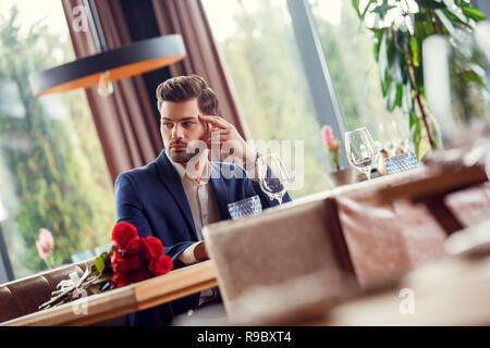 Young man on date in restaurant sitting at table with red rose bouquet looking aside touching face thinking serious Stock Photo