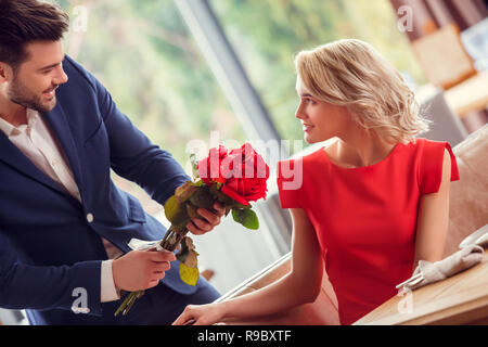 Young man and woman on date in restaurant guy standing smiling shy giving red roses bouquet to lady sitting at table looking at him surprised Stock Photo