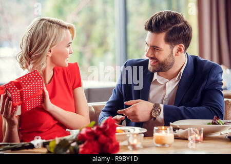 Young man and woman on date in restaurant sitting at table lady holding red present box looking at guy smiling playful Stock Photo