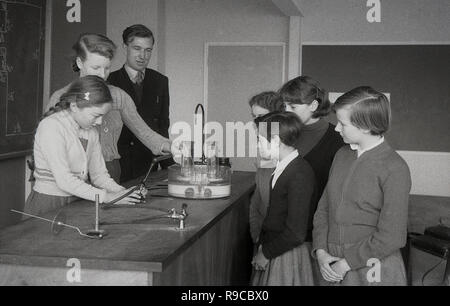 1955, a schoolgirl doing an experiment at a workbench in a science class, helped by a teacher and watched by her fellow pupils, England, UK Stock Photo